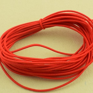 Puede incluir: Una bobina de cordón elástico rojo brillante, bien enrollada y anudada. El cordón es liso y parece estar hecho de un material sintético. El fondo es de color beige claro.
