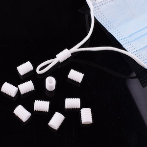 May include: White plastic cord stoppers for face masks. The stoppers are small, round, and have a hole in the center. They are arranged on a black surface.