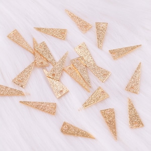 May include: A collection of gold-colored triangle-shaped metal charms with a hole at the top. The charms have a textured surface and are scattered on a white fuzzy background.