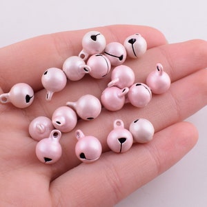 Peut inclure: Un ensemble de petites cloches en m&eacute;tal rose avec des clapets noirs. Les cloches sont &eacute;parpill&eacute;es sur une main et ont une finition m&eacute;tallique brillante.