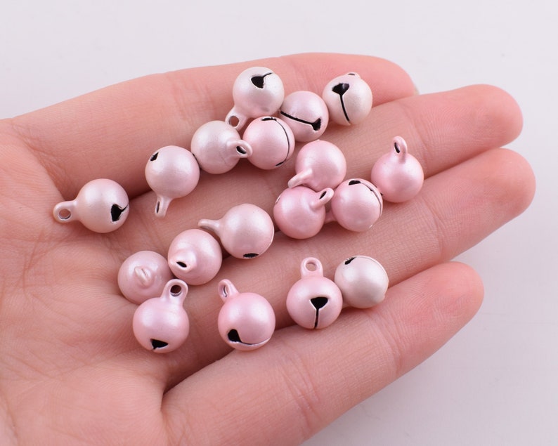 Peut inclure: Un ensemble de petites cloches en m&eacute;tal rose avec des clapets noirs. Les cloches sont &eacute;parpill&eacute;es sur une main et ont une finition m&eacute;tallique brillante.