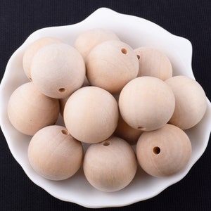 May include: A collection of unfinished, light-colored wooden beads, each with a drilled hole, displayed in a white, scalloped-edge bowl. The beads are spherical and appear smooth, perfect for crafting projects.