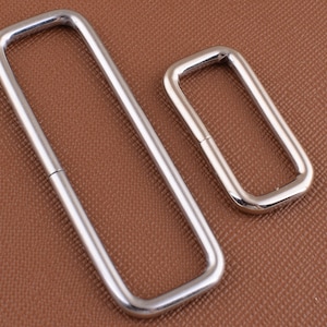 Könnte beinhalten: Zwei silberne D-Ringe aus Metall, einer größer als der andere, auf einer braunen strukturierten Oberfläche.