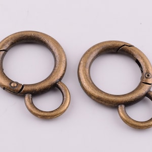 May include: Two antique bronze-colored spring snap rings. Each ring is circular with a hinged opening and a small, attached loop. The metal has a slightly aged appearance, suggesting a vintage or rustic style. These rings are suitable for various crafting and accessory projects.