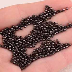 May include: A large pile of small, round, black beads. The beads are smooth and shiny, and they are scattered on a hand.