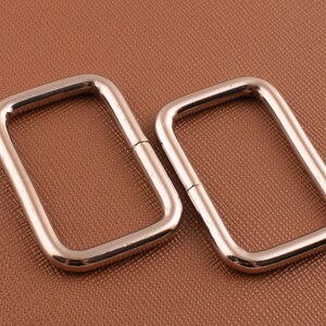 May include: Two silver metal D-rings, one slightly larger than the other, on a brown textured surface.