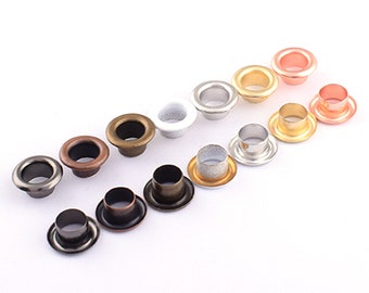 Op de afbeelding: Assortiment van metalen oogjes in verschillende afwerkingen, waaronder zilver, goud, brons en koper. De oogjes zijn in een rij op een witte achtergrond gerangschikt.