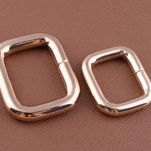 May include: Two gold-colored metal D-rings, one larger than the other, on a brown textured surface.