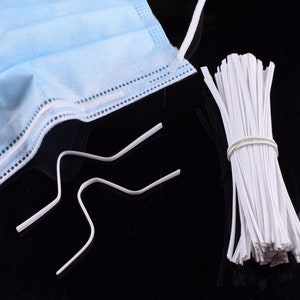 May include: A close-up of white plastic face mask nose wire and a bundle of white plastic face mask ear loops. The nose wire is bent into a U-shape.