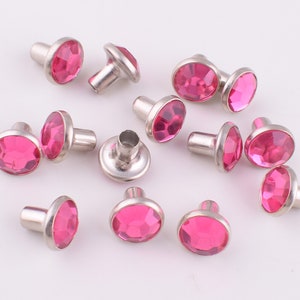 Op de afbeelding: Een verzameling roze strass studs met zilveren metalen bases. De studs zijn verspreid over een wit oppervlak.