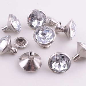 May include: Eight silver-toned, crystal-embellished, decorative knobs with a round, faceted crystal center. The knobs have a flat base and a raised, rounded top.
