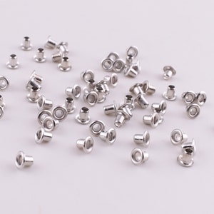 May include: A large quantity of silver metal eyelets, also known as grommets, scattered on a white surface. These eyelets are used for reinforcing holes in fabric and other materials.