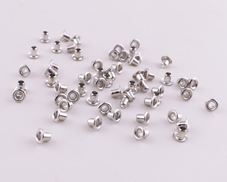May include: A large quantity of silver metal eyelets, also known as grommets, scattered on a white surface. These eyelets are used for reinforcing holes in fabric and other materials.
