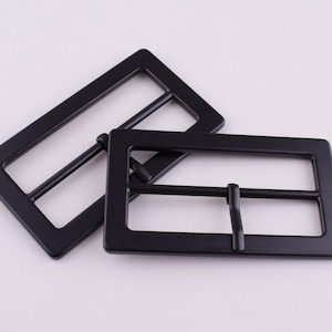 Puede incluir: Dos hebillas de cinturón de metal negro. Las hebillas son rectangulares con un acabado liso y mate.