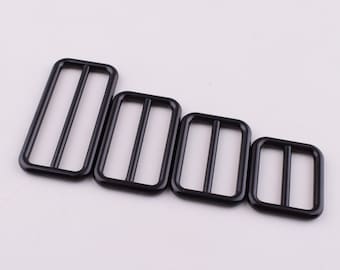 May include: Four black metal slider buckles of different sizes. The buckles are rectangular with two parallel bars in the centre.