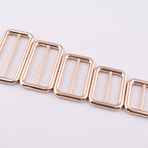 May include: Five gold-toned metal slider buckles of varying sizes. The buckles are rectangular with a smooth finish and a single, centered opening.