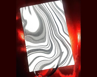 Könnte beinhalten: Ein rechteckiges Notizbuch mit einem schwarz-weißen Wirbelmuster auf dem Einband. Das Notizbuch hat ein schwarzes Lesebändchen. Der Hintergrund ist eine glänzende rote Oberfläche.