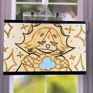 May include: A drawing of a cartoon character with blonde hair, a blue flower-like shape, and a sad expression. The artwork is on a cream-colored background with gold accents and is held by a clear clip.