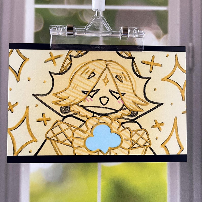 May include: A drawing of a cartoon character with blonde hair, a blue flower-like shape, and a sad expression. The artwork is on a cream-colored background with gold accents and is held by a clear clip.