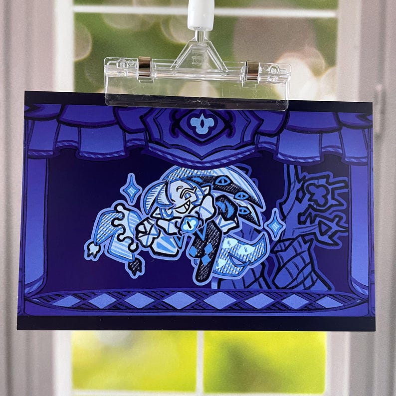 May include: A blue and white illustration featuring a cartoon character on a stage with a curtain backdrop. The character has a playful expression and is surrounded by stylized elements. The artwork is held by a clear plastic clip.