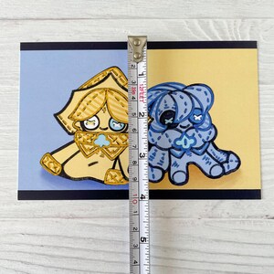 May include: A postcard featuring two cartoon characters, one gold and one blue, each with stitched details. The gold character is on a blue background, and the blue character is on a yellow background. The postcard measures approximately 4 inches in height.