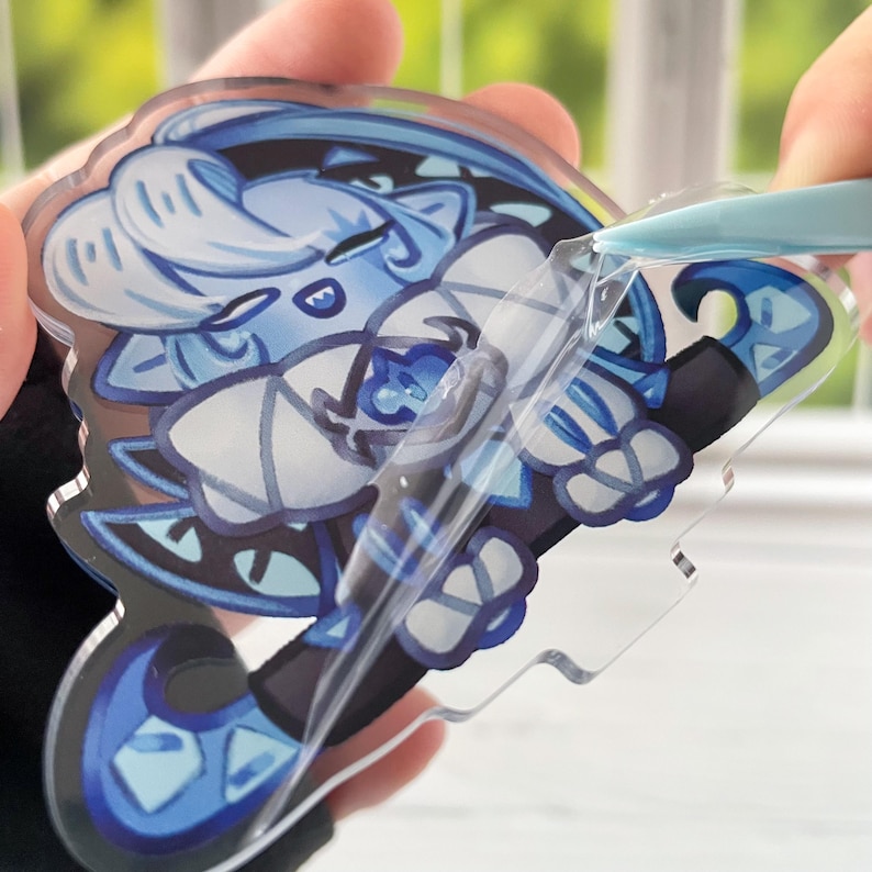 K&ouml;nnte beinhalten: Ein klarer Acryl-Anh&auml;nger mit einem blau-wei&szlig;en Cartoon-Charakter-Design. Der Charakter hat ein stilisiertes Aussehen mit blauen Haaren und Kleidung. Ein Werkzeug l&ouml;st eine Schutzschicht ab und gibt die gl&auml;nzende Oberfl&auml;che des Anh&auml;ngers frei.
