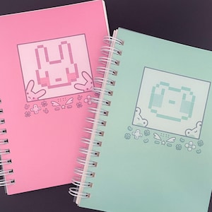 May include: Two spiral bound notebooks with pink and green covers. The pink notebook has a pixelated bunny rabbit on the front and the green notebook has a pixelated frog on the front.