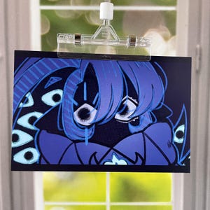 May include: A rectangular art print featuring a stylized blue character with multiple eyes and blue hair. The artwork is held by a clear plastic clip, with a window and greenery visible in the background.