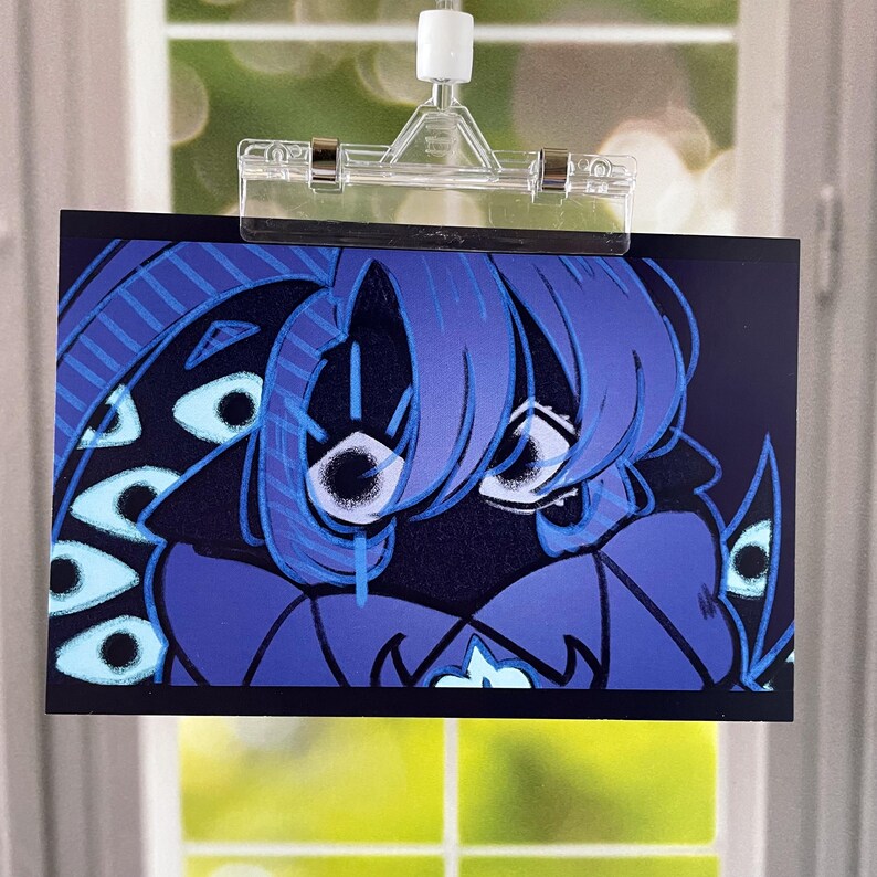 May include: A rectangular art print featuring a stylized blue character with multiple eyes and blue hair. The artwork is held by a clear plastic clip, with a window and greenery visible in the background.