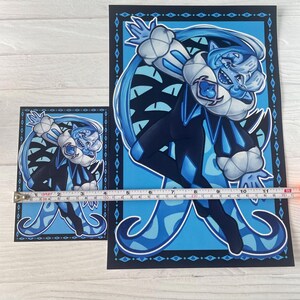 May include: Two art prints featuring a blue cartoon character with a white collar and a diamond pattern on the clothing. The larger print measures approximately 28 cm in height. The background is light blue with a dark blue border.