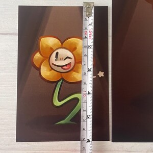 May include: A yellow flower with a smiling face and a red tongue. The flower has a green stem and is on a brown background.