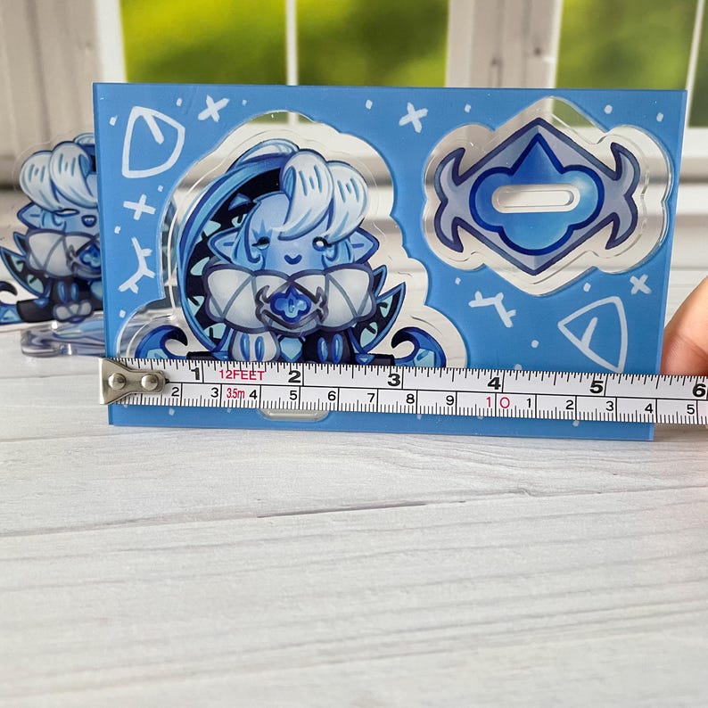 K&ouml;nnte beinhalten: Ein blauer Acryl-Anh&auml;nger mit einer Cartoon-Figur mit hellblauen Haaren und Akzenten. Der Anh&auml;nger ist etwa 12,7 cm breit. Das Design umfasst einen dekorativen Rand mit wei&szlig;en Akzenten und Formen.