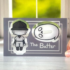 Puede incluir: Un expositor de acrílico plateado con una ilustración en blanco y negro de un jugador de béisbol y un fantasma. El texto "The Batter" está impreso en el expositor.