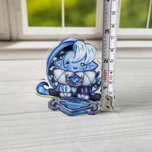 K&ouml;nnte beinhalten: Ein klarer Acryl-Anh&auml;nger mit einer blau-wei&szlig;en Cartoon-Figur. Der Anh&auml;nger ist etwa 7.6 cm hoch und hat einen dekorativen Sockel. Die Figur hat gro&szlig;e Augen und einen freundlichen Ausdruck. Das Design ist stilisiert.