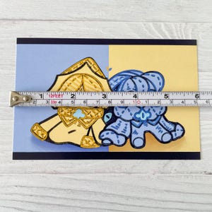 May include: A postcard featuring two cartoon characters, one yellow and one blue, with a tape measure across the image. The tape measure shows a length of approximately 5 inches.