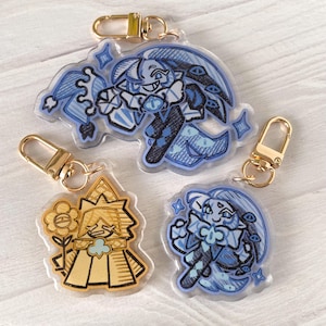 May include: Three acrylic keychains with gold-colored clasps. Two keychains feature blue and black cartoon characters, while the third has a gold and black cartoon character. The keychains are on a light-colored surface.