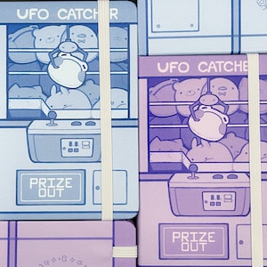 May include: Two purple and blue notebooks with the text "UFO Catcher" and "Prize Out" on the front. The notebooks have a cartoon design of a claw machine with stuffed animals inside.