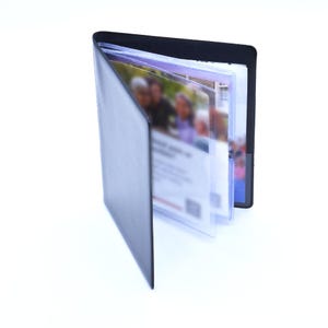 May include: A black leatherette business card holder, open to display several clear plastic sleeves filled with cards. The holder is rectangular with rounded corners and a vertical orientation. The background is white.