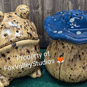 May include: Two ceramic figurines, a frog and a mushroom, both with brown speckled glaze. The frog is light brown with dark brown spots and the mushroom is blue with white spots. The mushroom has a brown stem with a small orange fox figurine on it. The text "Property of FoxValleyStudios" is visible on the frog.