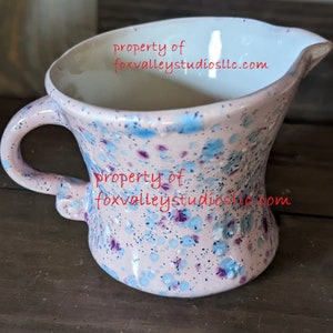 May include: A pink ceramic pitcher with a blue and purple speckled glaze. The pitcher has a handle and a spout. The text "property of foxvalleystudiosllc.com" is visible on the pitcher.