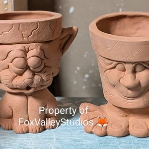 May include: Two terracotta planter pots with whimsical faces. One pot is shaped like a cat with closed eyes and a small nose. The other pot is shaped like a person with a smiling face and closed eyes. The pots are both unglazed and have a rough texture. The text "Property of FoxValleyStudios" is visible on the bottom of the image.