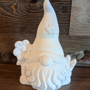 May include: White ceramic gnome figurine with a pointed hat and a beard. The gnome has a flower on its hat and another flower on its shoulder.