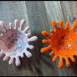 May include: Two ceramic bowls with a splatter paint design. One bowl is pink with blue and purple speckles, the other is orange with yellow and blue speckles.