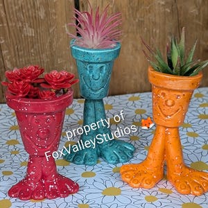 May include: Three whimsical, colorful planters with smiling faces and foot-shaped bases. The planters are red, teal, and orange, each holding a different succulent. The red planter has white speckles. The text "property of FoxValleyStudios" is visible.