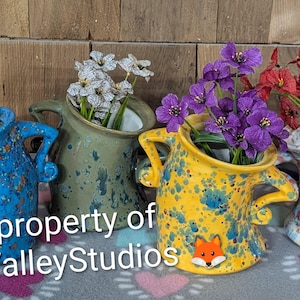 May include: Four ceramic pitchers with two handles each, featuring speckled glaze in blue, olive green, yellow, and white. Each pitcher contains artificial flowers in shades of pink, white, purple, and red. A small fox icon is on the yellow pitcher.