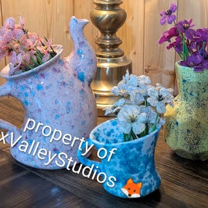May include: A collection of ceramic vases and pitchers in various shapes and sizes. The largest is a pink pitcher with blue speckles, holding pink flowers. A blue cup with white flowers and a yellow vase with purple flowers are also present. The text "property of FoxValleyStudios" is visible.