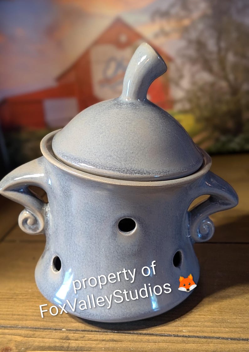 May include: A pale blue ceramic oil warmer with a lid and three holes. The warmer has two handles with a curled design. The text "property of FoxValleyStudios" is visible on the front.