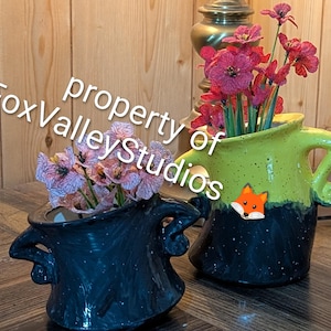 May include: Two ceramic pitchers with handles, one solid dark blue and one two-toned lime green and dark blue, both featuring white speckles. The pitchers contain artificial flowers in shades of pink and red. Text reads property of FoxValleyStudios.