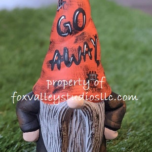 May include: A small orange gnome with a black and white painted hat that says "GO AWAY". The gnome is wearing a black outfit and has a white beard.