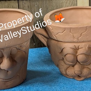 May include: Two terracotta-colored face planters with detailed facial features. One planter has a smiling face, and the other has a cat-like face with ears. A small fox figurine is inside one of the planters. The text "Property of FoxValleyStudios" is visible.